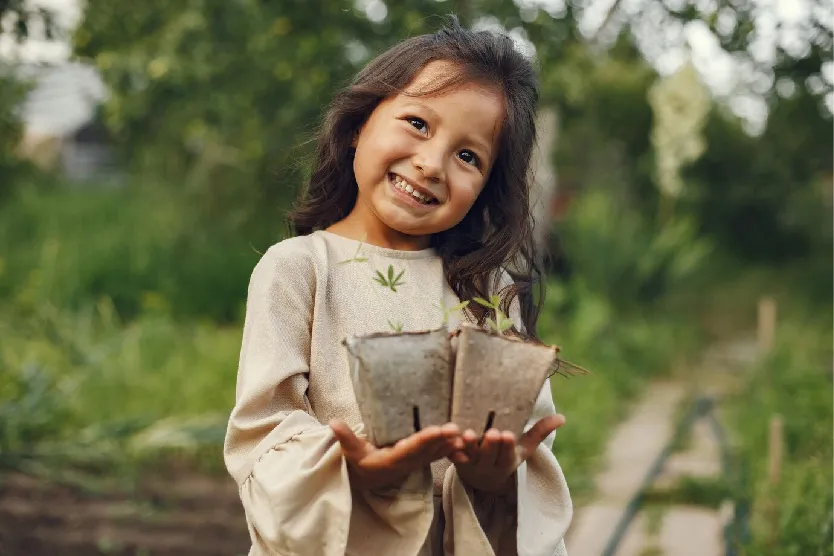 Foto de una niña sonriendo, sosteniendo con sus dos manos dos macetas de flores